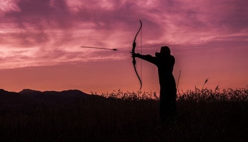 young woman shooting bow at sunset like katniss everdeen